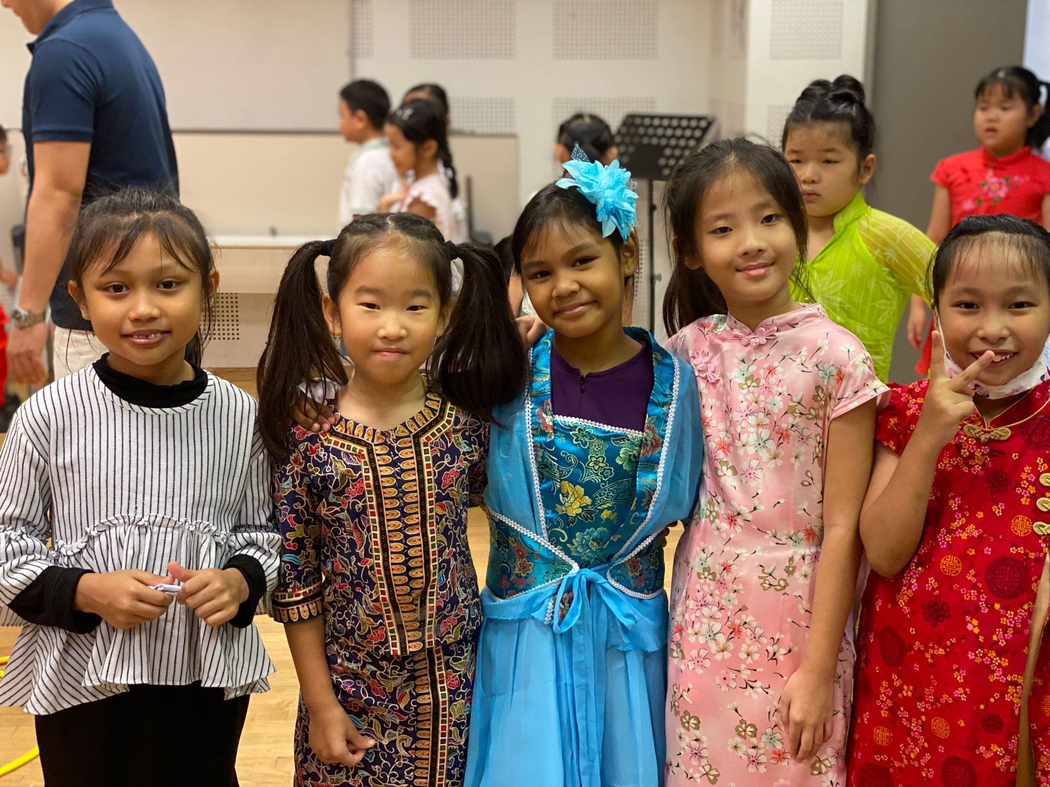 Five young girls smile together wearing colorful traditional Asian outfits including cheongsams and batik dress indoors.